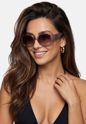 Kobieta z długimi brązowymi włosami w dużych brązowych okularach przeciwsłonecznych, złotych kolczykach kołach i czarnej bluzce, uśmiechająca się i dotykająca obojczyka.