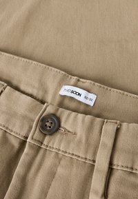 Pantaloni beige in cotone con superficie strutturata, dotati di un bottone marrone, un'etichetta in vita con la scritta "Two Soon" e taglia 32/32.
