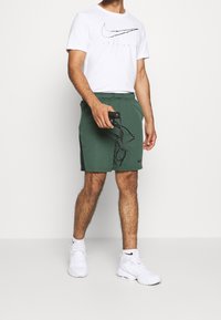 Pantalones cortos deportivos verdes con detalles laterales negros, combinados con una camiseta blanca que lleva un logo negro de Nike y zapatillas blancas.