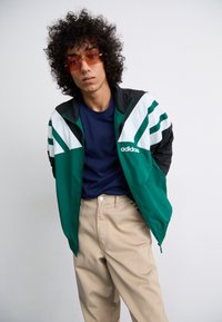 Green and black track jacket with white stripes, logo on chest. Worn over a blue crewneck shirt, paired with beige trousers.