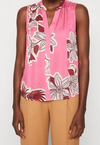 Rosa ärmlös blus med blommönster i rött och vitt. Har ett urtagande krage och en knapp vid halslinningen. Mjuk, slät tyg.