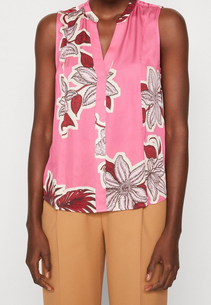 Rosa ärmlös blus med blommönster i rött och vitt. Har ett urtagande krage och en knapp vid halslinningen. Mjuk, slät tyg.