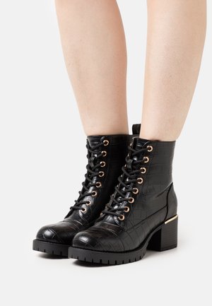 Botas de cuero negro texturizadas hasta el tobillo con cordones, ojales dorados y tacones en bloque, llevadas sobre piernas descubiertas contra un fondo blanco.