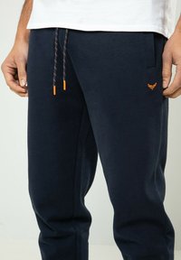 Homme portant un pantalon de survêtement bleu marine avec embouts de cordon orange et petit logo brodé orange sur la cuisse gauche, associé à une chemise blanche.