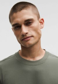 Camicetta grigio-verde a maniche corte con scollatura rotonda, realizzata in tessuto morbido, con dettagli sottili sulle cuciture delle spalle e una consistenza liscia.