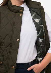 Nahaufnahme einer Person in einer olivgrünen gesteppte Weste mit einem Fasanen-Musterfutter, einem weißen Hemd und einer Hand, die in der Tasche einer dunklen Jeans steckt.