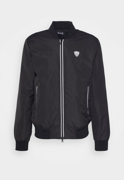 Veste aviateur noire en tissu lisse, dotée d'une fermeture éclair, d'accents blancs, de poches latérales et d'un écusson logo sur la poitrine gauche.