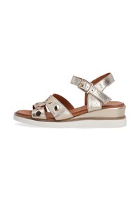 Metallic guld sandaler med ankelremme, dekorative nitter og kilehæle på en hvid baggrund. Velegnet til stilfuld sommerbeklædning.