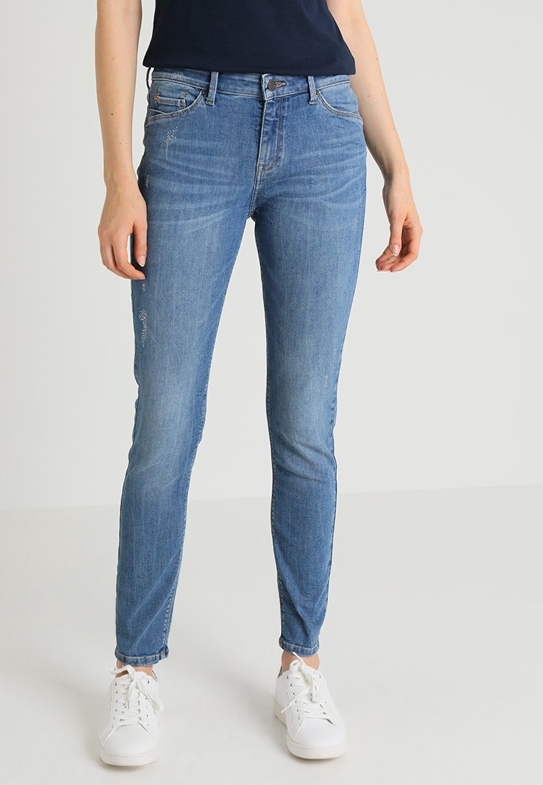 Femme portant un jean bleu slim et des baskets blanches, debout devant un fond gris clair uni.