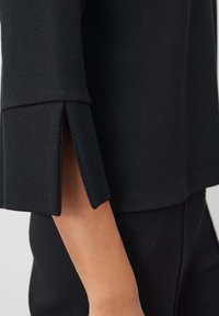 Haut noir en tissu à texture lisse avec détail fendu sur les manches, présentant un design simple et épuré.