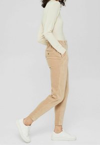 Kvinna i beiga kordflorsbyxor, krämfärgad långärmad topp, vita sneakers, som går med händerna i fickorna mot en enfärgad bakgrund.