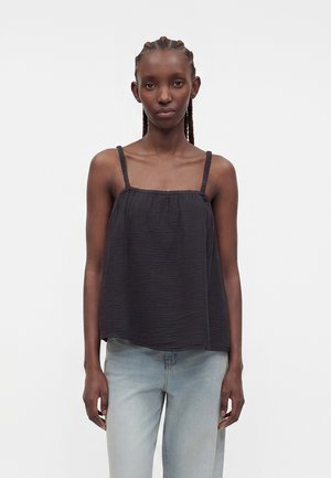 Γυναίκα με πλεγμένα μαλλιά που φοράει φαρδιά μαύρη αμάνικη μπλούζα και ανοιχτό μπλε τζιν, στέκεται μπροστά σε απλό λευκό φόντο.