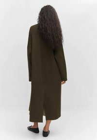 Frau mit langen lockigen Haaren, die einen langen olivgrünen Mantel mit einem Schlitz hinten und schwarzen flachen Schuhen trägt, vor einem einfarbigen Hintergrund stehend.