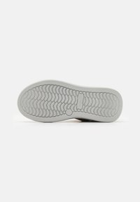 Suela de zapato con una base de goma texturizada y un patrón de banda de rodadura en zigzag, color gris claro y un logotipo destacado en el centro.