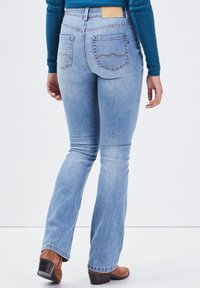 Jasnoniebieskie dzwony z wysokim stanem, pięcioma kieszeniami i brązową skórzaną metką z tyłu. Materiał ma gładką fakturę.