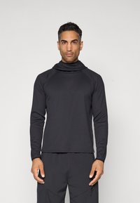 Svart termisk langermet topp med funnel neck, med en teksturert ribbestruktur og kontraststing, båret over mørke bukser.