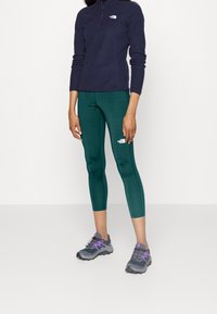 Turkusowe legginsy, szare buty sportowe z fioletowymi akcentami i granatowa bluza z rozpinanym suwakiem do 1/4. Legginsy mająlogo oraz miękką fakturę.