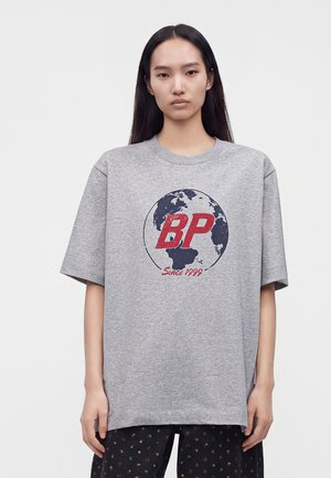 Femme aux longs cheveux foncés portant un t-shirt gris oversize avec un motif de globe et des lettres rouges "BP Since 1999", debout devant un fond clair uni.