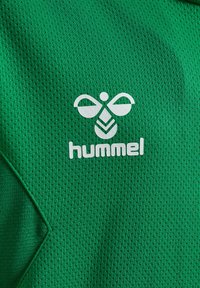 Grønt tekstureret stof med et diamantmønster, forsynet med et centralt hvidt trykt logo af to vinger og en dråbe, under ordet "hummel."