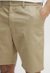 Beige Shorts aus leichtem Baumwollstoff, mit lockerer Passform, Fronttaschen und einem Knopfverschluss am Bund. Glatte Textur.