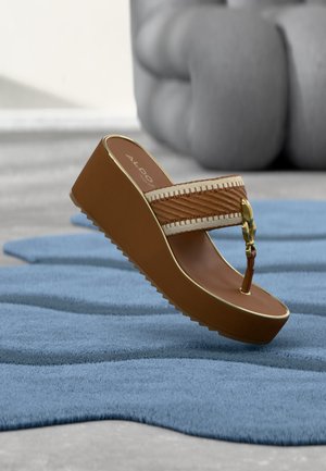 Sandale à talon compensé marron avec bride tissée beige et détail boucle dorée, posée sur un tapis bleu texturé avec un canapé gris flou en arrière-plan.