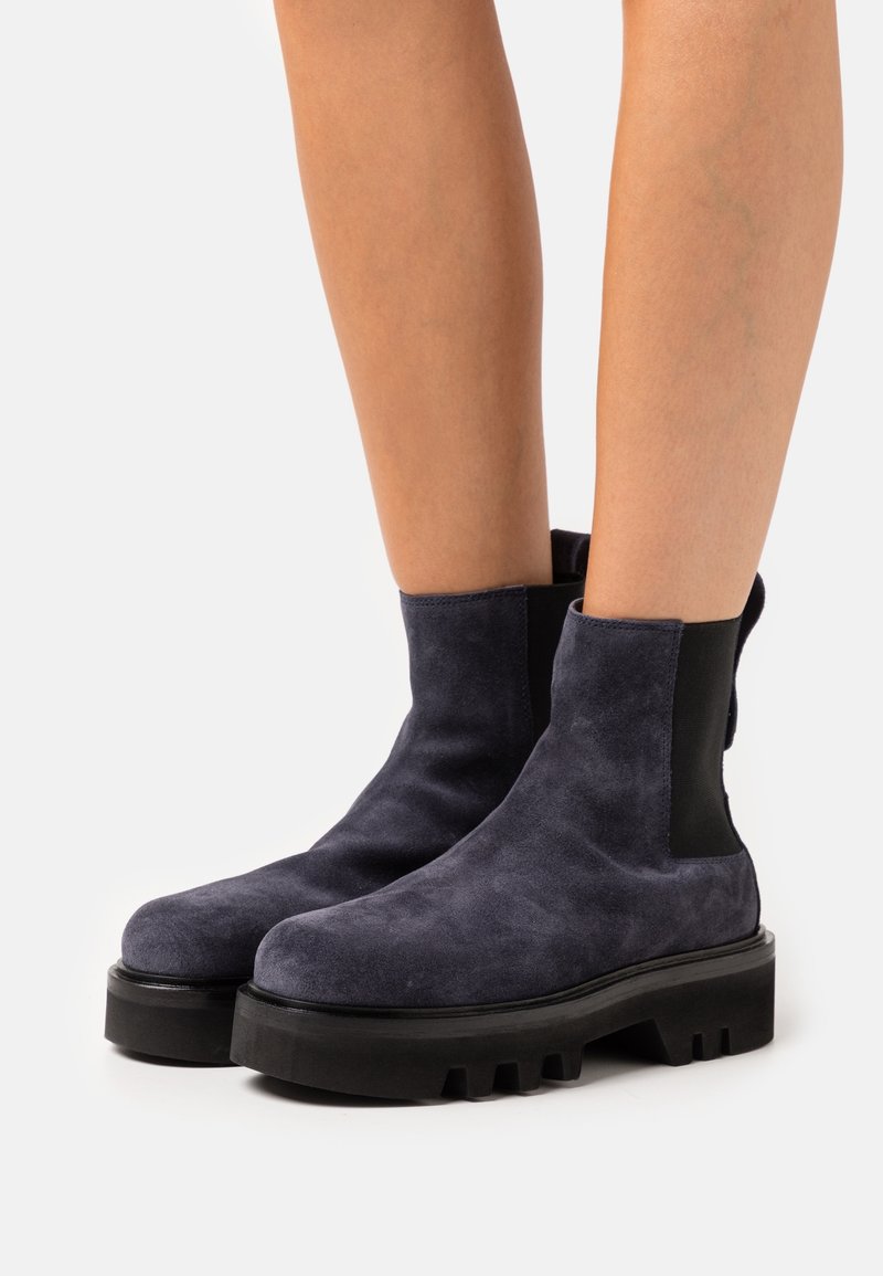 Jambes portant des bottines en daim bleu marine avec des semelles carrées noires épaisses et des panneaux latéraux élastiques sur un fond blanc uni.