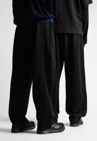 Dva človeka, oblečena v črne široke hlače z belim obrobjem in črnimi usnjenimi čevlji, stojita drug ob drugem na beli podlagi.