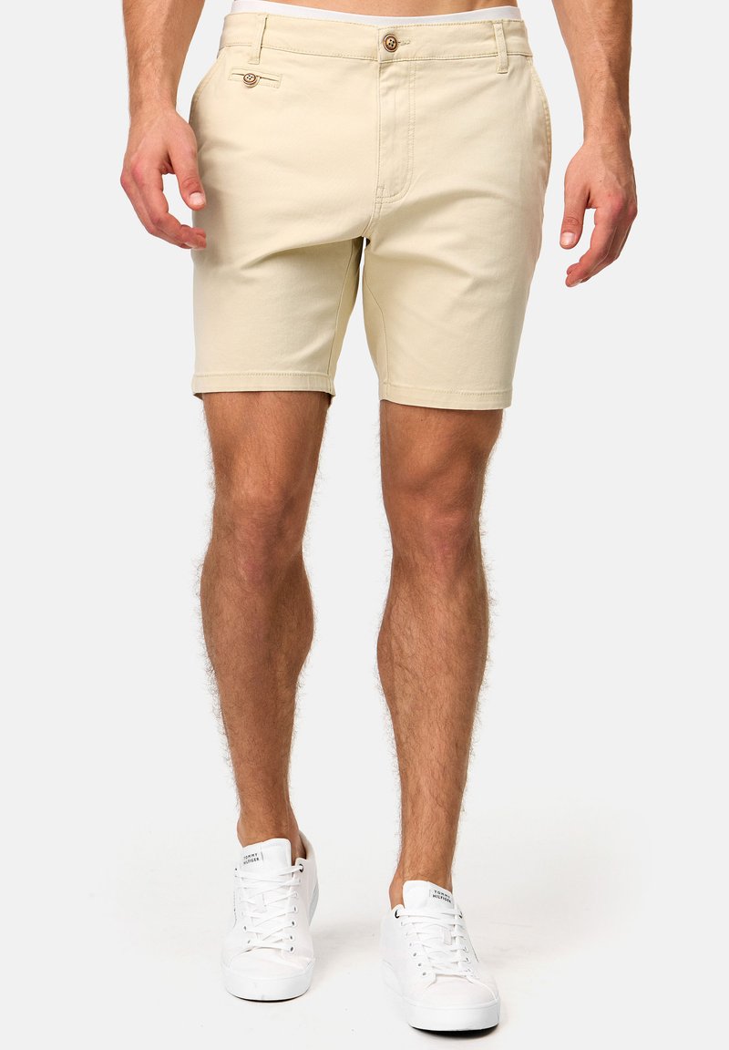 Pantalones cortos de algodón beige con un frente plano, dos bolsillos laterales y cierre de botón. Presenta un dobladillo enrollado y un diseño clásico. Combinados con zapatillas blancas.