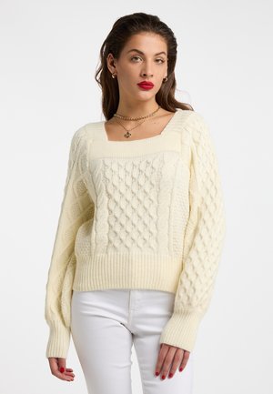 Donna che indossa un maglione crema a trecce con scollo quadrato, abbinato a pantaloni bianchi e rossetto rosso.