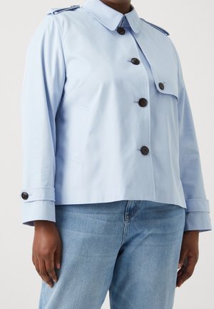 Person trägt eine hellblaue Jacke mit Knöpfen und Schulterklappen sowie hellblaue Jeans und steht vor einem schlichten Hintergrund.