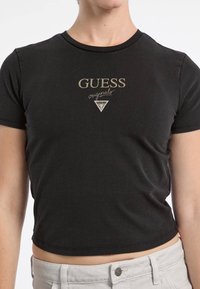 Czarny krótki t-shirt z okrągłym dekoltem, z metalicznym złotym napisem "GUESS originals" oraz trójkątnym detalem logo na klatce piersiowej.