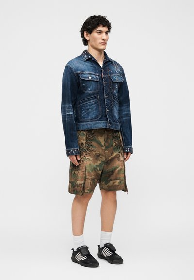 Νέος άνδρας που στέκεται φορώντας ένα μπλε τζιν μπουφάν με διακοσμητικά, καμουφλάζ cargo σορτς, λευκές κάλτσες και μαύρα αθλητικά παπούτσια.