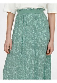 Kvinde iført en grøn blomsterkjole med elastisk talje, parret med en hvid t-shirt med korte ærmer, hænderne afslappet ned langs siden.