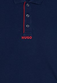 Námořnická polo košile vyrobená z texturované tkaniny s polo zapínáním na tři knoflíky. Obsahuje červený kontrastní límec a červené logo "HUGO" na hrudi.