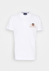 Bílá polo košile vyrobená z bavlny s klasickým límečkem, obsahující knoflíkovou légu a vyšívané logo v červené a zlaté barvě na hrudi.