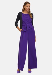 Lila overáll széles lábbal, megkötős derékrésszel és testhez simuló felső résszel. Sötét textúrájú kardigánnal és azonos lila cipővel párosítva.