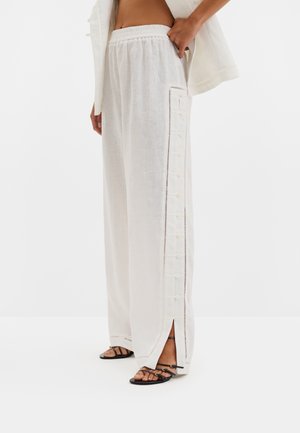 Pantalon blanc à jambes larges avec boutons latéraux et petites fentes aux chevilles, porté avec des sandales plates noires à lanières.