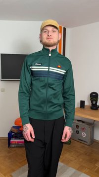 Grön zip-up jacka med vita och mörkblå horisontella ränder, med en logotyp på bröstet. Bärs med svarta byxor.