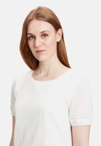 Weißes gestricktes Oberteil mit kurzen Ärmeln, das ein Lückenmuster aufweist, sowie einen gerippten Rundhalsausschnitt und eine glatte Textur. Zu den Details gehören runde Aussparungen im gesamten Design.