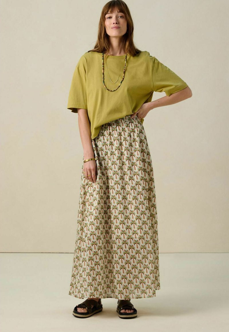 T-shirt en coton surdimensionné olive associé à une longue jupe crème ornée d'un motif de feuilles de palmier en vert et rose, et des sandales noires.