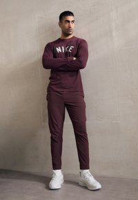 Bordo garais piedurkņu T-krekls ar "NIKE" logo, pārī ar atbilstošām sporta biksēm un baltām sporta kurpēm. Gluda auduma, pieguļošs dizains.