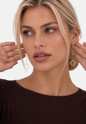 Femme blonde portant de longues boucles d’oreilles en chaîne dorée avec des pendentifs circulaires, touchant ses cheveux, regardant légèrement sur le côté sur un fond uni.