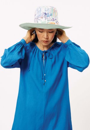 Blauwe jurk met lange mouwen, een trekkoorddecolleté en knoopdetails, gecombineerd met een hoed met brede rand en een kleurrijk patroon.