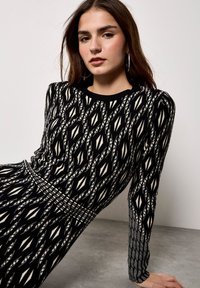 Czarna i biała sukienka w wzory z okrągłym dekoltem, długimi rękawami i dopasowanym krojem. Charakteryzuje się geometrycznym wzorem i teksturowanym dzianym materiałem.