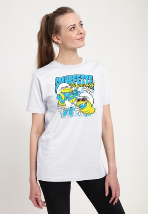 Junge Frau in einem weißen Smurfette-T-Shirt mit blauen Smurfette-Figuren und dem Text „Sei du selbst“, stehend vor einem schlichten Hintergrund.