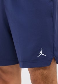 Short de sport bleu marine sur la cuisse d’une personne affichant un logo Jumpman blanc près de l’ourlet avec une petite fente latérale.