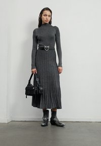 Maglione grigio lavorato a maglia con maniche lunghe abbinato a una gonna midi plissettata grigia. Include una cintura nera con una fibbia circolare argentata e stivaletti.