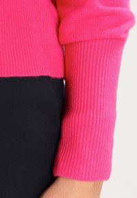 Strickpullover in einem lebhaften Pinkton, mit geripptem Textur an den Ärmelbündchen, kontrastiert mit einem glatten schwarzen Stoff darunter.