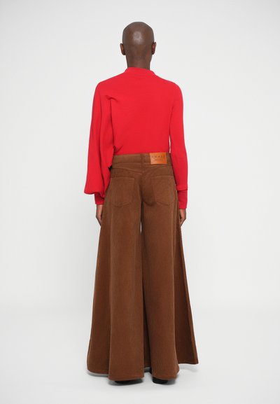 Camiseta roja ajustada de manga larga combinada con pantalones de pana marrones de pierna ancha, que presentan bolsillos traseros y una etiqueta en la cinturilla.