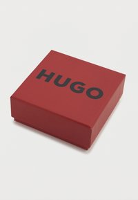 Κόκκινο ορθογώνιο κουτί με απαλή υφή, με τη λέξη "HUGO" σε έντονους μαύρους κεφαλαίους χαρακτήρες στην κορυφή.